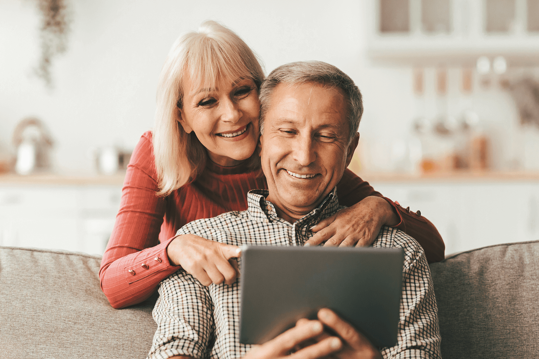 Älteres Paar sitzt auf dem Sofa mit Tablet und lacht, Sinnbild für Geborgenheit und Komfort im eigenen Haus