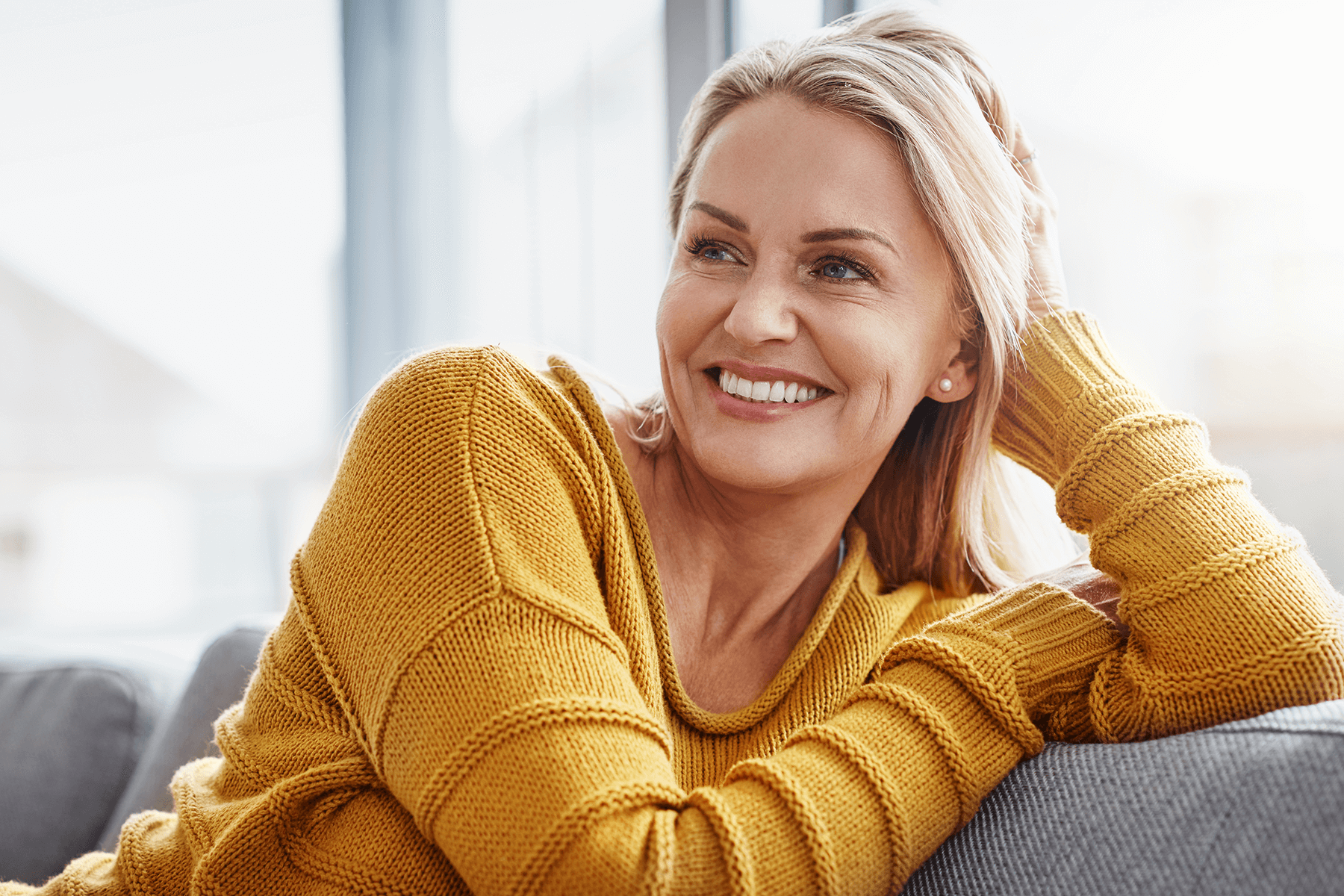 Frau mit blondem Haar lehnt entspannt am Fenster und lächelt, Ausdruck von Zufriedenheit und Wohlbefinden im neuen Zuhause