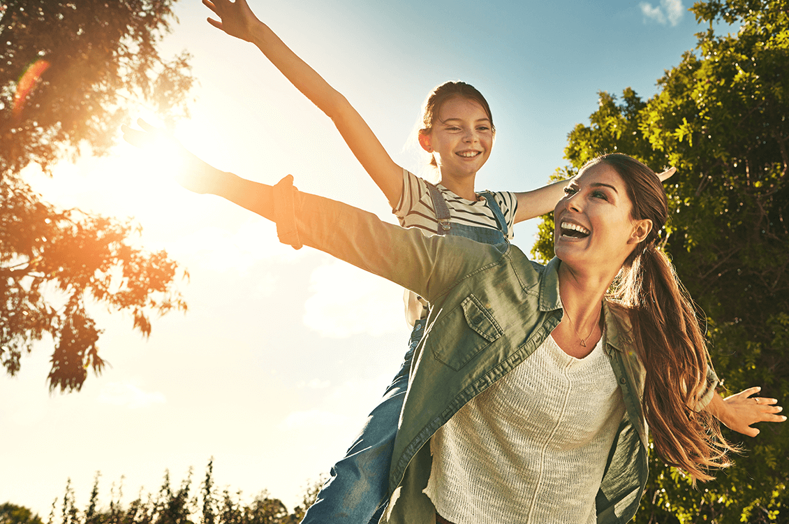 Frau im Garten wedelt mit einem Arm glücklich. Mit der anderen Hand hält sich ihre Tochter hinter dem Rücken die freudig in den Sonnenstrahlen zusammen mit ihrer Mutter im Garten eines Vionda Bungalows rennt.