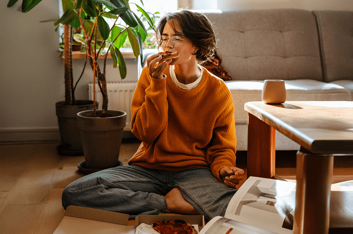 Person sitzt entspannt auf dem Boden im Wohnzimmer, isst ein Stück Pizza, warmes Licht, Sofa und Zimmerpflanzen im Hintergrund