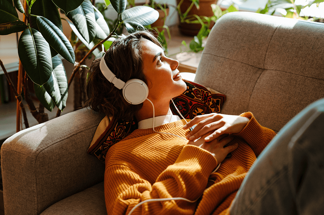 Person liegt entspannt auf dem Sofa im Wohnzimmer, hört Musik mit Kopfhörern, warmes Tageslicht und grüne Pflanzen sorgen für ruhige Atmosphäre