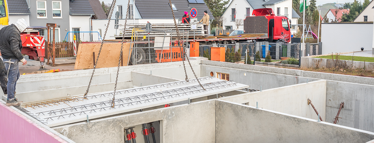 Baustelle mit montierten Kellerwänden; Betondeckenelemente werden mit Ketten in Position gehoben.