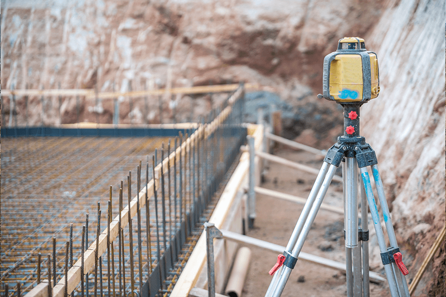 Baustelle mit vorbereiteter Bodenplatte und Bewehrungsstahl; im Vordergrund steht ein Vermessungsgerät auf einem Stativ.