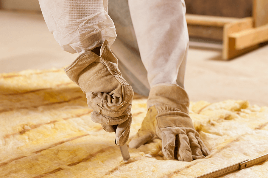 Handwerker mit Arbeitshandschuhen schneidet Dämmmaterial aus Mineralwolle auf dem Boden während einer Sanierungsmaßnahme.