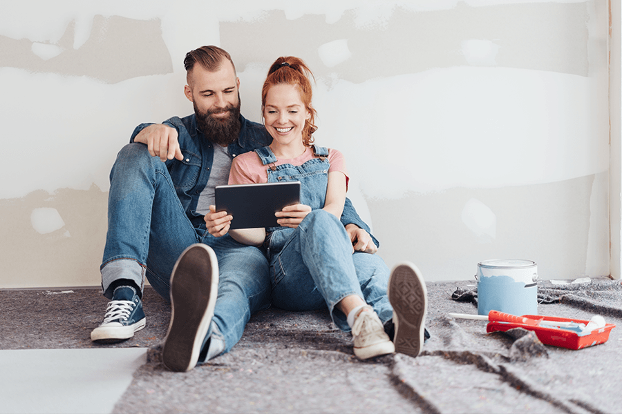 Paar sitzt auf dem Boden einer Baustelle in einer Wohnung, umgeben von Werkzeug und Farbeimern, und schaut gemeinsam auf ein Tablet.