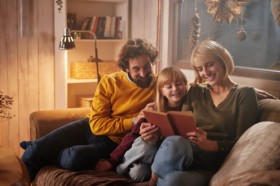 Familie sitzt gemeinsam auf einem Sofa und liest entspannt in einem warm beleuchteten Wohnzimmer